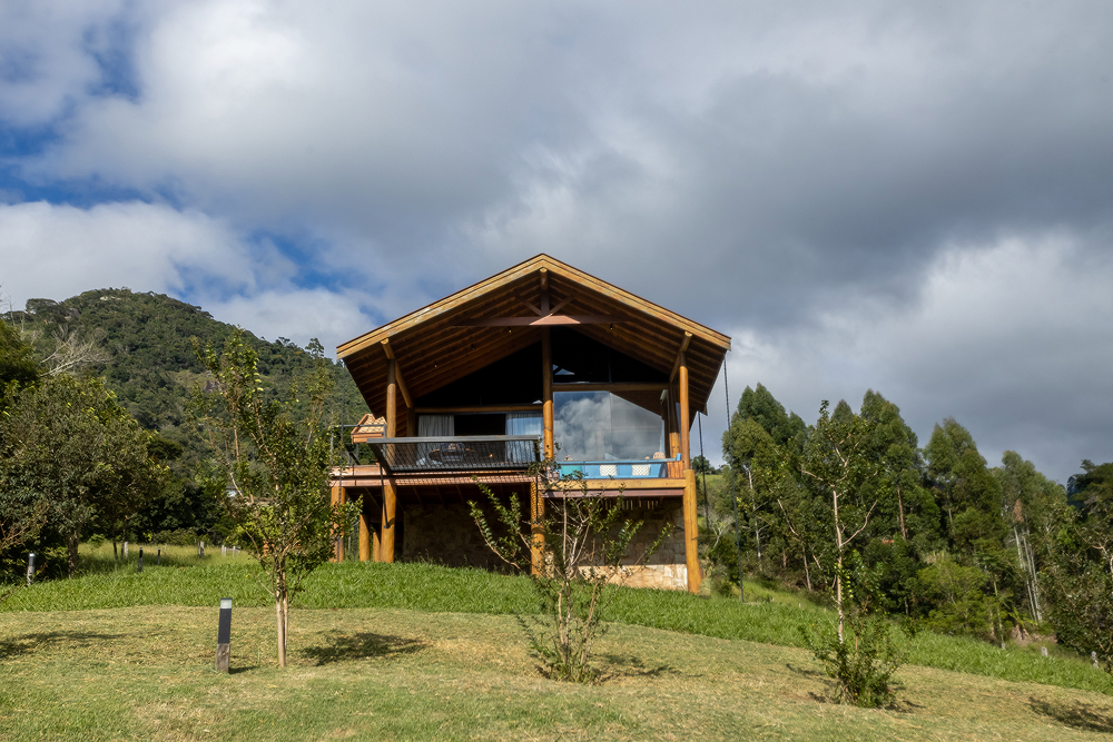 Stone and wood cabin on a grassy hill with a large tree beside it under a bright sun and blue sky with wispy clouds.