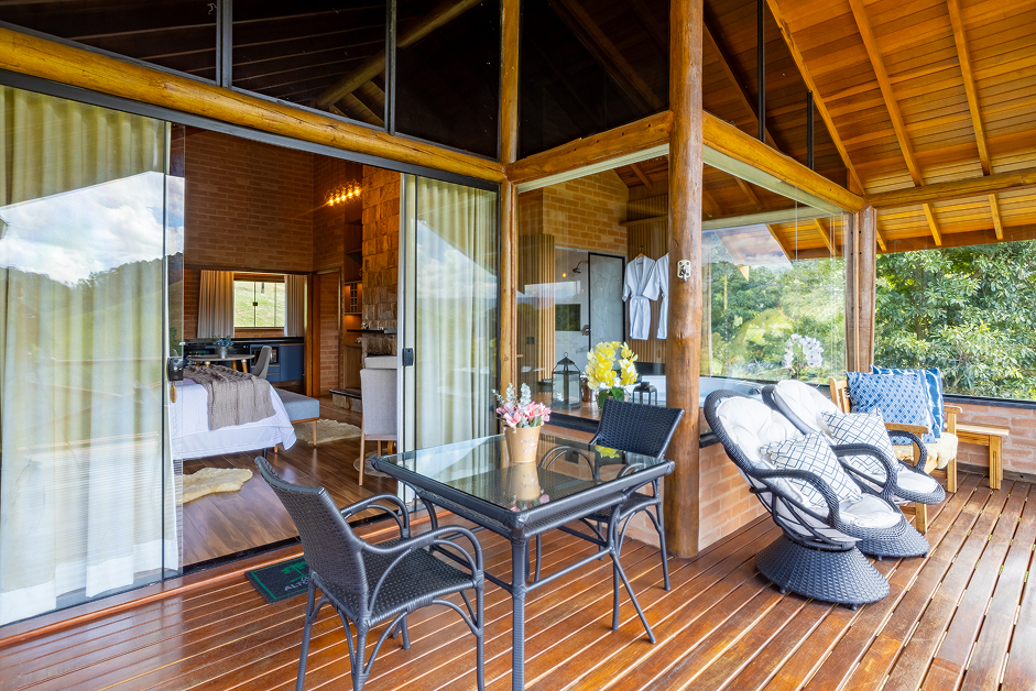 Wooden porch with glass table, black wicker chairs, cushioned swivel chairs, and view into a cozy bedroom and living area through large glass doors.