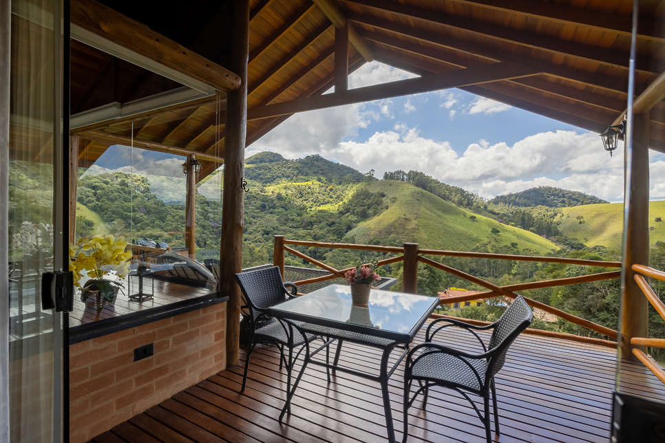 Wooden deck with black chair and table next to a cabin, overlooking a lush green mountainous landscape with clear sky and a large yellow plant in the foreground.