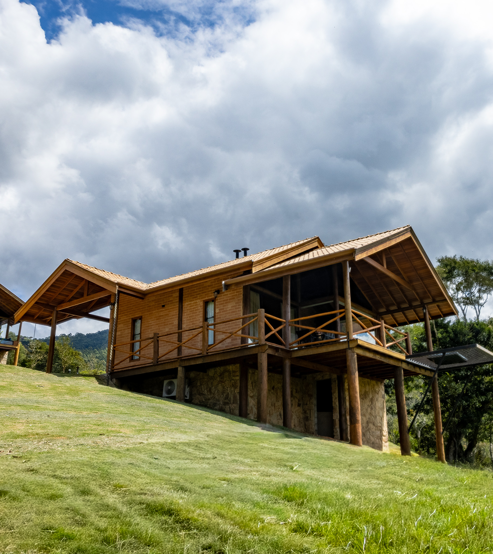 Stone and wood cabin on a grassy hill with a large tree beside it under a bright sun and blue sky with wispy clouds.