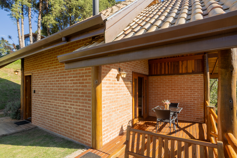 Wooden swing bench with pergola on a wooden deck next to a brick house with a tiled roof under a blue sky.