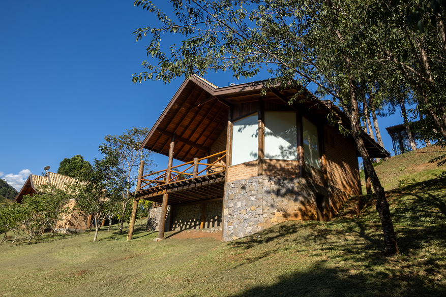 Wooden deck with black chair and table next to a cabin, overlooking a lush green mountainous landscape with clear sky and a large yellow plant in the foreground.