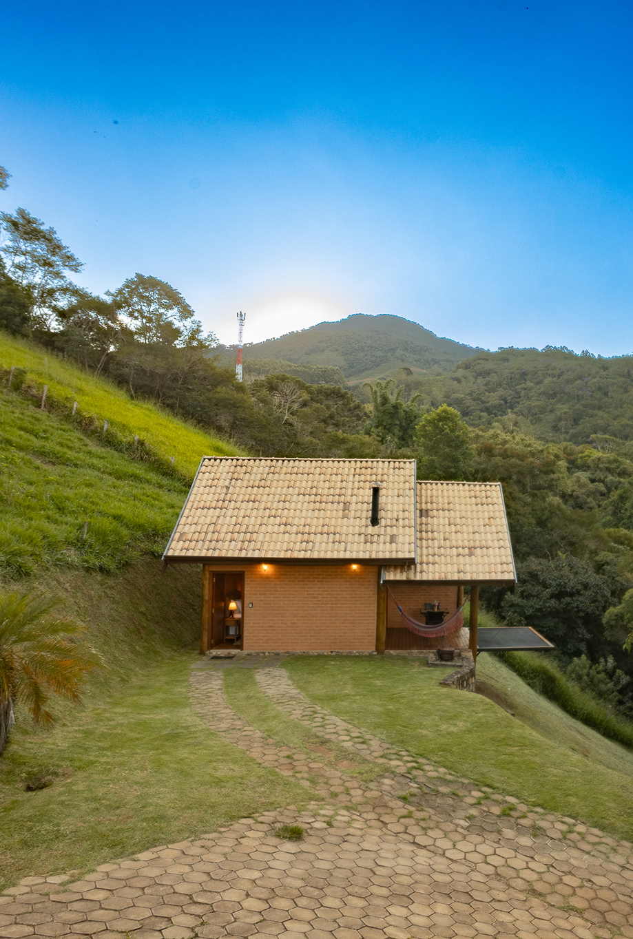 Stone and wood cabin on a grassy hill with a large tree beside it under a bright sun and blue sky with wispy clouds.