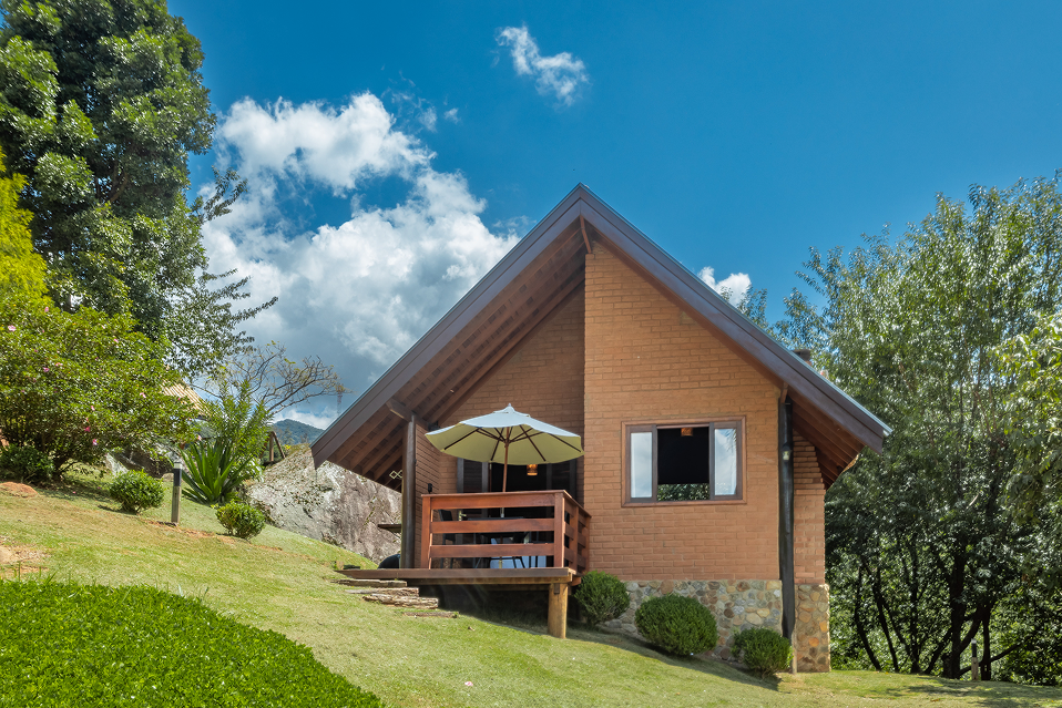 Wooden swing bench with pergola on a wooden deck next to a brick house with a tiled roof under a blue sky.