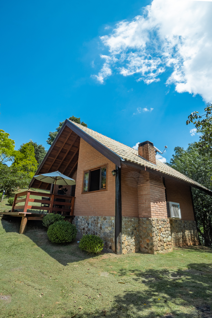 Stone and wood cabin on a grassy hill with a large tree beside it under a bright sun and blue sky with wispy clouds.