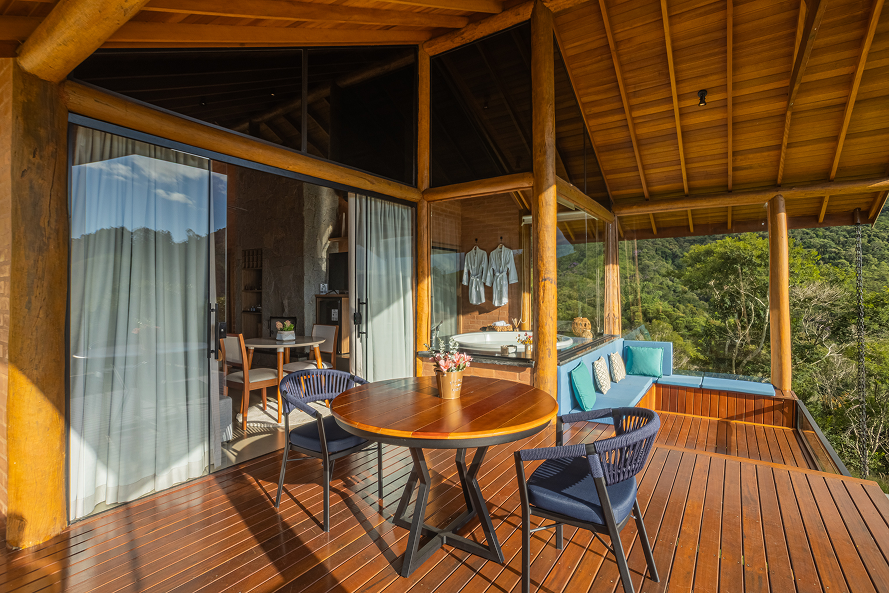 Modern wooden house on stilts with large glass windows, surrounded by grassy lawn and mountains under a clear blue sky.